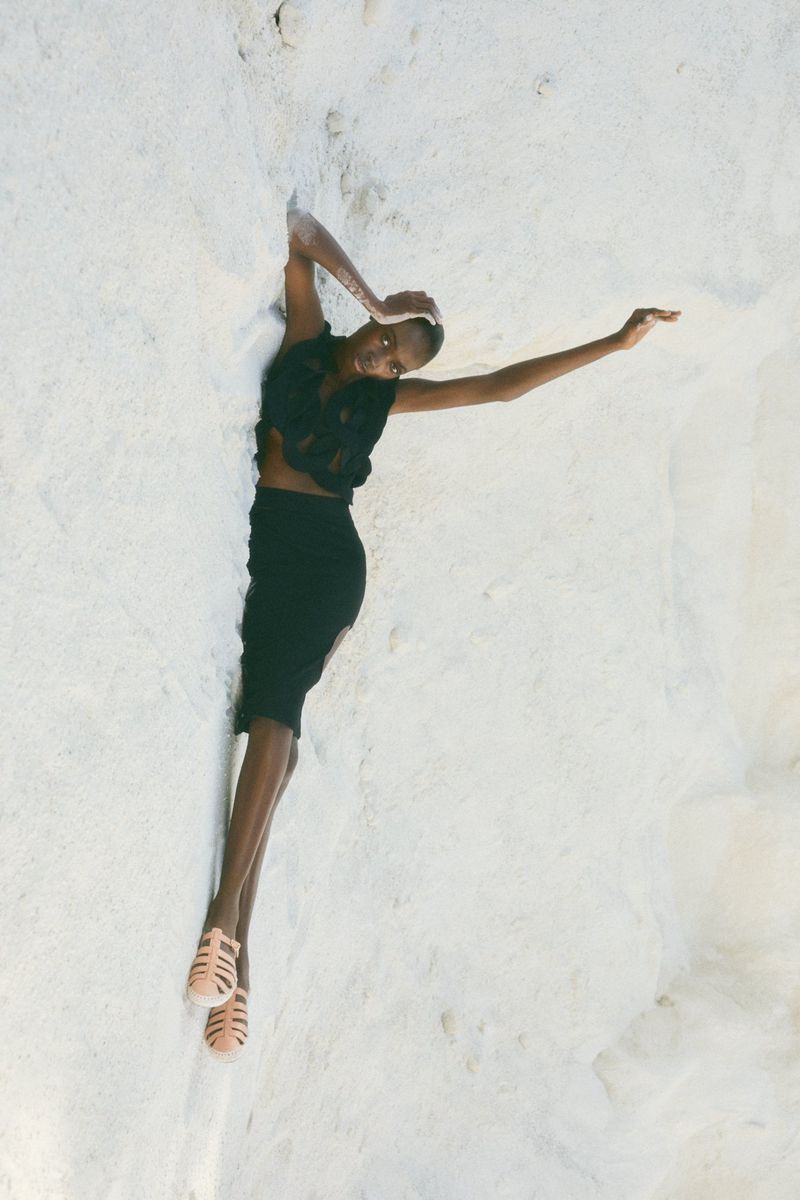 A woman in a black outfit lying on a white textured surface, posing with outstretched arms. The scene features soft lighting.