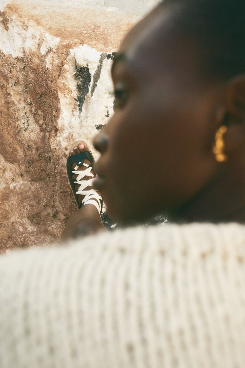 Close-up portrait of a Black model wearing laced sandals against a rocky coastal background, photographed by Lala Serrano.