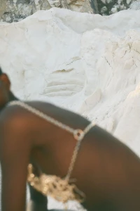 Model wearing gold chain body harness posed against a textured white stone quarry background for Ancient Greek Sandals x Castaner.