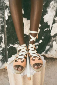 Model Janelle Ononye wearing tan leather lace-up espadrille sandals standing on a fluted stone pedestal.