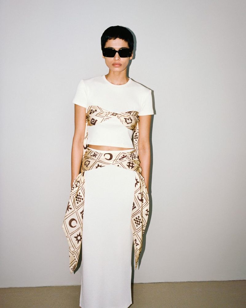 A woman with short dark hair and sunglasses, wearing a white skirt and shirt with a patterned sash, stands against a gray backdrop.
