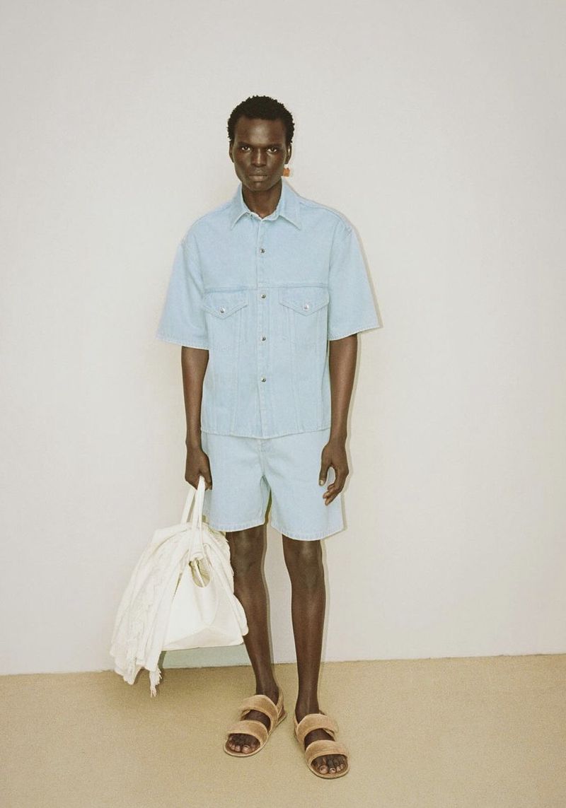 A dark-skinned male model poses in a matching denim shirt and shorts, holding a bag. He is wearing sandals against a neutral backdrop.