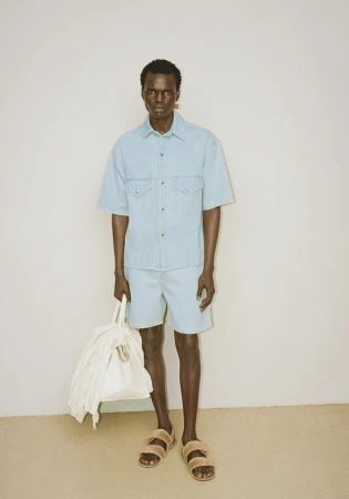 A dark-skinned male model poses in a matching denim shirt and shorts, holding a bag. He is wearing sandals against a neutral backdrop.