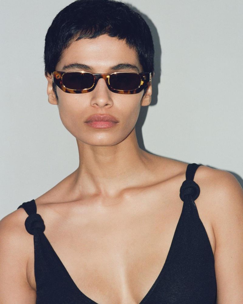 Close-up of a woman with short hair wearing tortoise-shell sunglasses and a black top against a neutral background, fashion studio shot.
