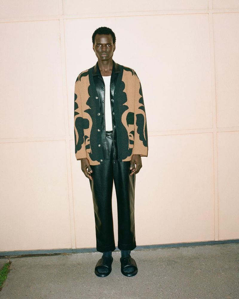 An adult man in a patterned shirt, black leather pants, and black slides poses in front of a pale peach wall on a concrete surface.