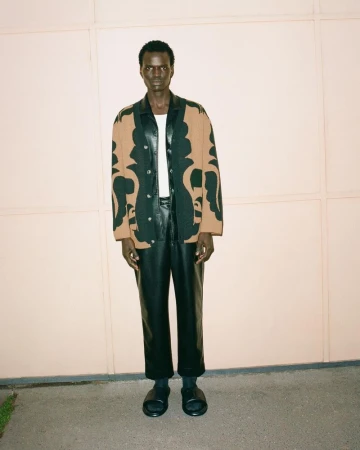 An adult man in a patterned shirt, black leather pants, and black slides poses in front of a pale peach wall on a concrete surface.