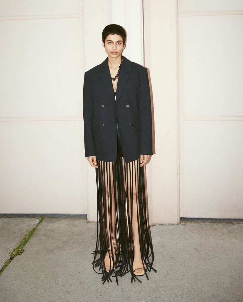 A woman poses in a studio, wearing a black blazer and a skirt made of long black fringe. The image is a fashion editorial shot.