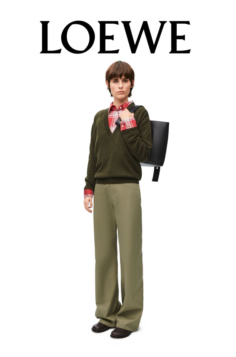 Fashionable adult female in an olive V-neck sweater and khaki pants holds a black tote bag against a white backdrop with 'LOEWE' above.