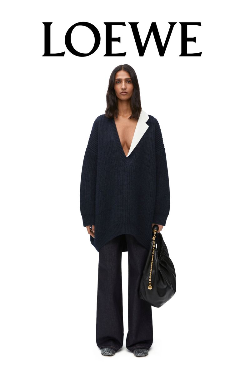 Fashion shot of a woman in a navy blue, oversized sweater and dark pants with a black purse in front of a white backdrop.