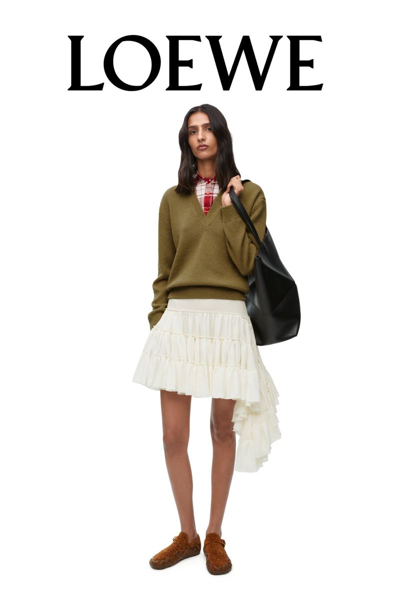 Model standing against a white backdrop wearing an olive green v-neck sweater, white ruffled asymmetric skirt, and large black shoulder bag.