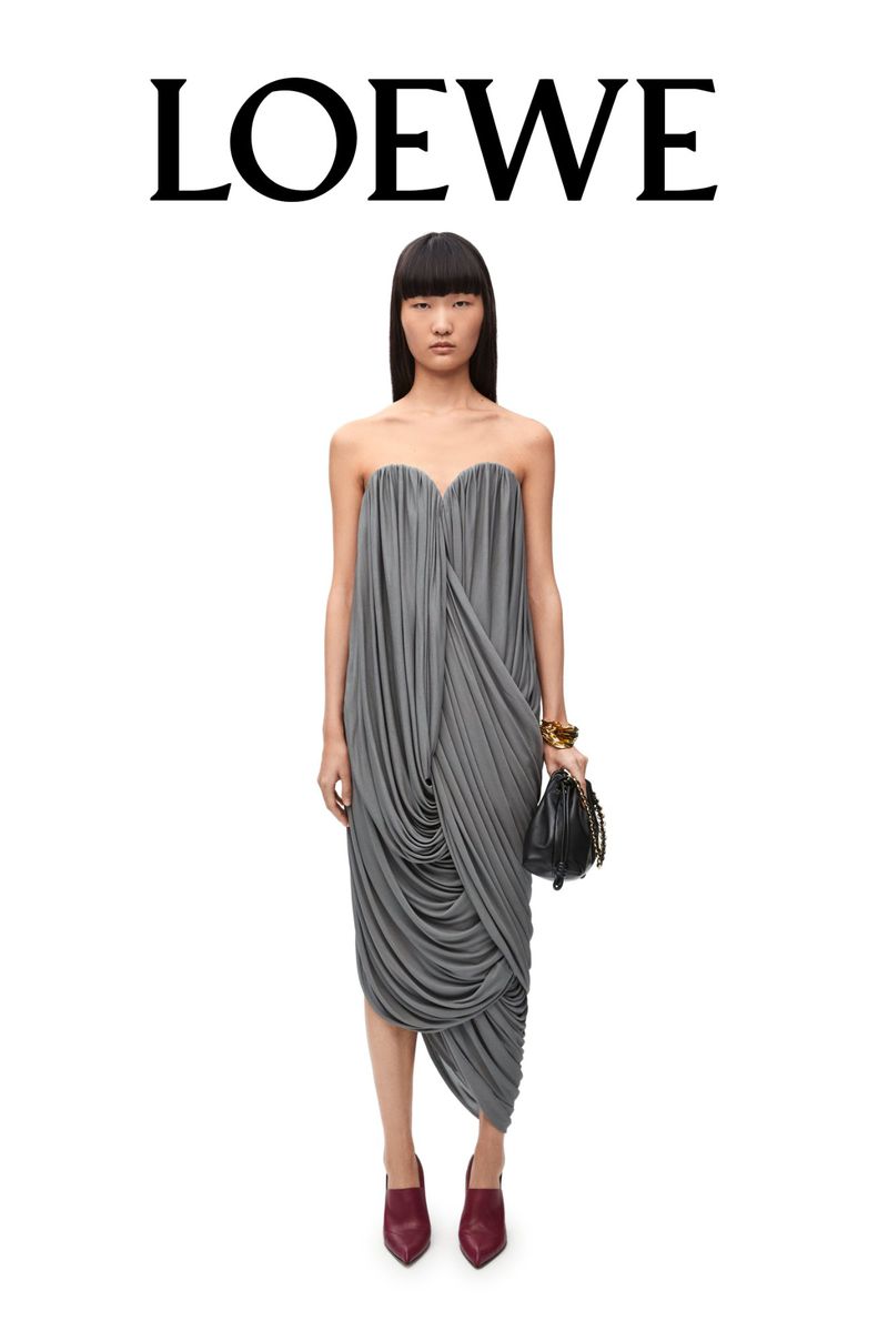 An Asian model poses in a grey draped dress with a black handbag, standing against a white backdrop with the LOEWE logo overhead.