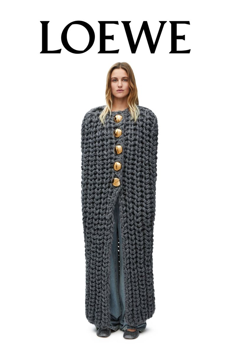 Woman wearing gray, oversized knitwear coat with large wooden buttons and wide leg pants in a fashion studio shot on a white backdrop.