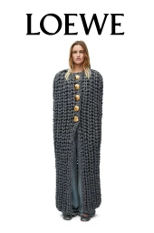 Model wearing a floor-length oversized grey chunky knit cape with large gold buttons against a white studio background.