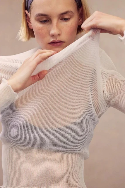A fair-skinned young adult female in a see-through top holds the fabric up with both hands in a neutral indoor setting.