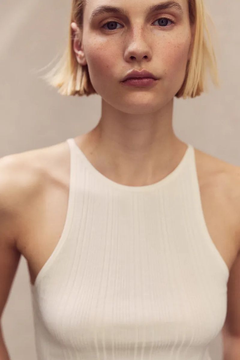 Close-up of an adult female with short blonde hair looking directly at the camera, wearing a light-colored ribbed tank top against a neutral background.