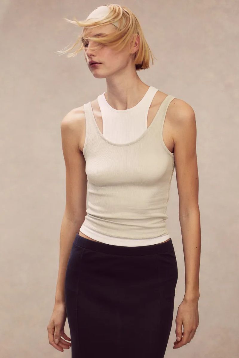 Woman in a studio setting wearing a beige tank top and a black skirt posing in front of a neutral background with soft lighting.