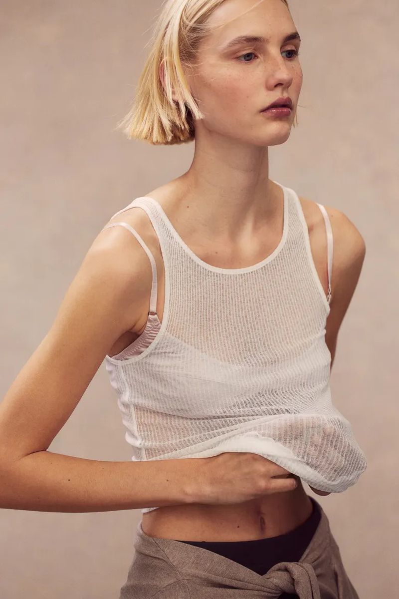 Model Elisa Nijman wears a semi-sheer white knit tank top lifted at the waist against a neutral warm studio backdrop for H&M.