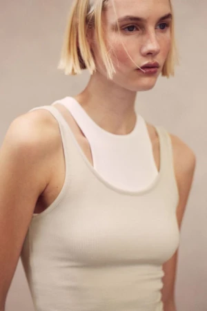 Model Elisa Nijman wearing layered white and off-white ribbed tank tops against a neutral studio backdrop.
