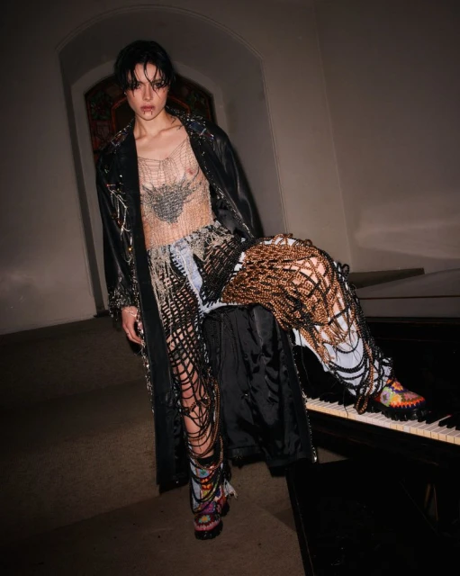 Fashionable young adult posing in avant-garde denim outfit, fishnet top, and long coat in an indoor setting with piano keys.