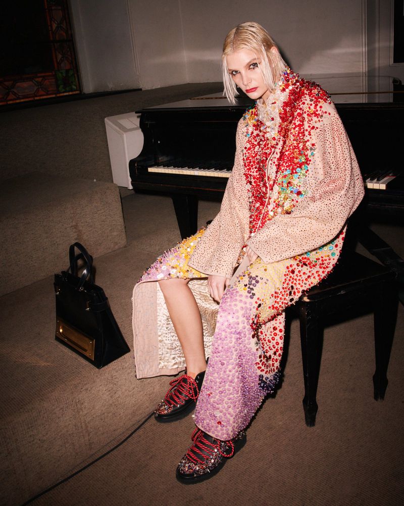 Fashion model sits on piano bench wearing patterned coat and sneakers with a handbag, shot in a dimly lit room with beige carpet.