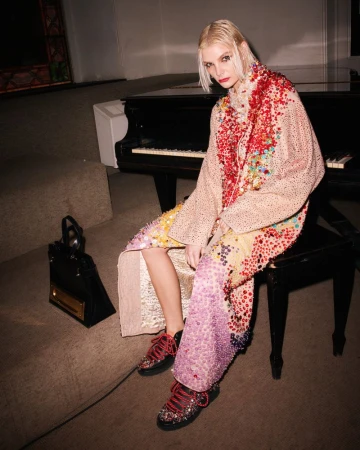 Fashion model sits on piano bench wearing patterned coat and sneakers with a handbag, shot in a dimly lit room with beige carpet.