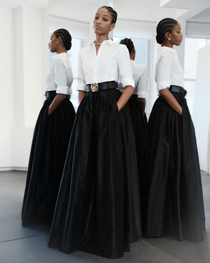 Elegant Black woman in long black skirt and white blouse poses with her hands in her pockets, reflected in mirrors in studio.