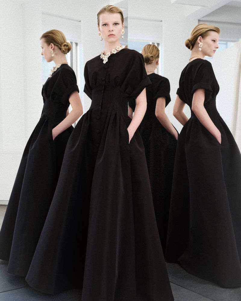 Woman in a black gown posing for a fashion shot, dress has an A-line skirt and short sleeves, accessorized with a pearl necklace and earrings.