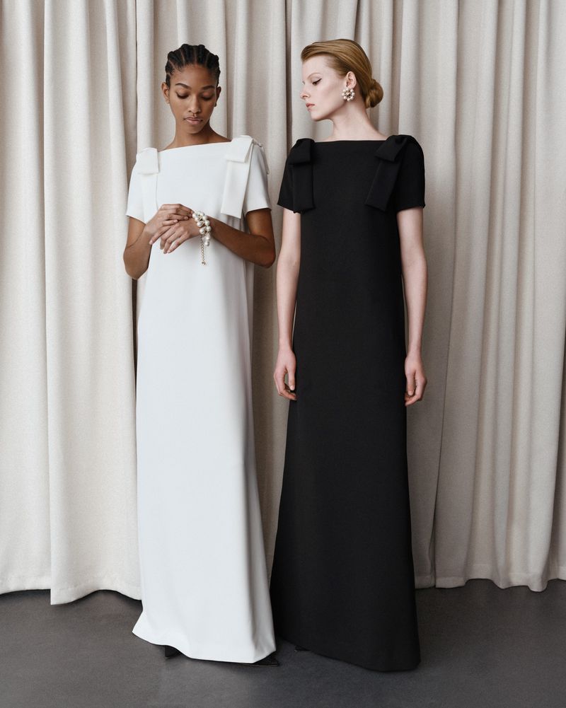 Two women in floor-length gowns, one in white with a pearl bracelet and the other in black, stand against a neutral curtain backdrop.