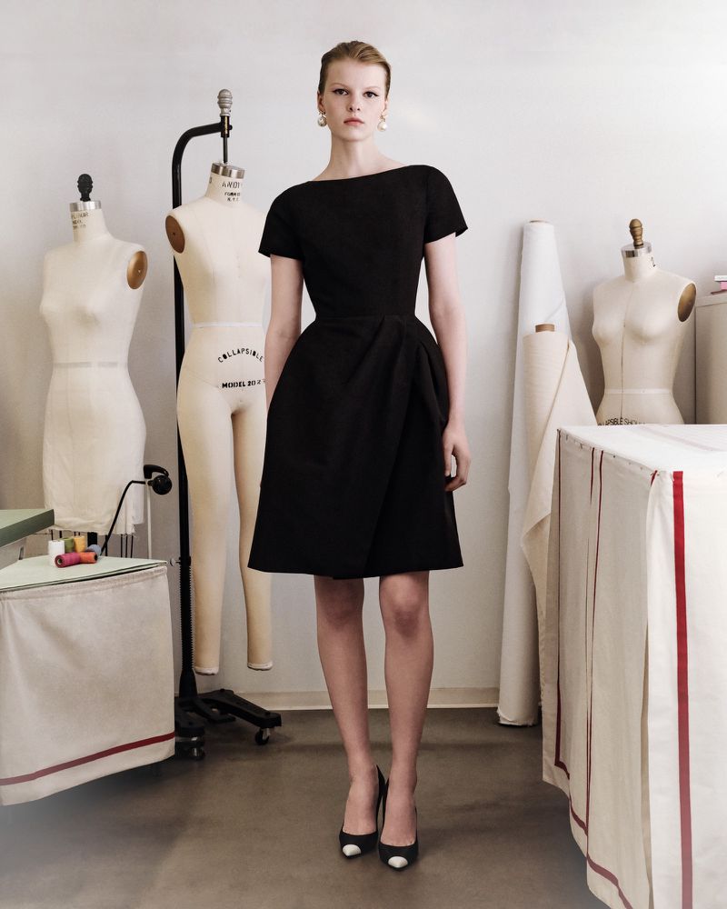Woman in a short black dress and black and white heels stands in a bright studio with dress forms in the background.
