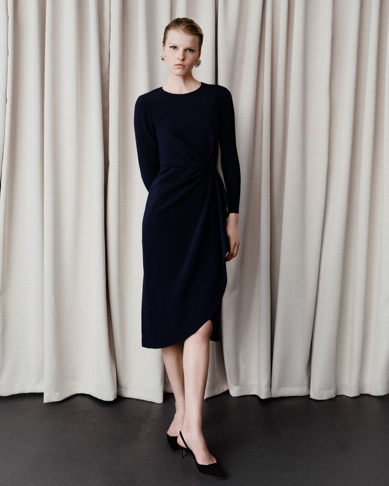 A slender woman poses in a navy midi dress with draped detailing against a neutral curtain backdrop in a studio setting.