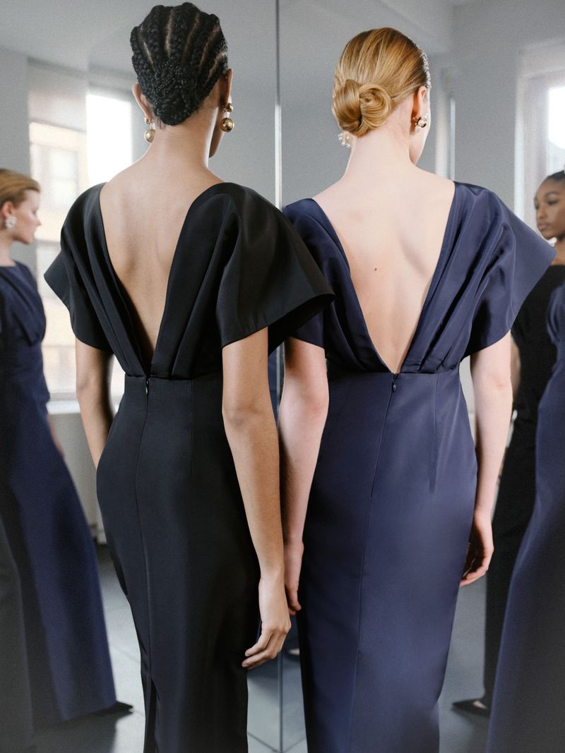Two women in elegant, dark open-back dresses are captured indoors with a neutral background, reflected in a mirror with two other women.