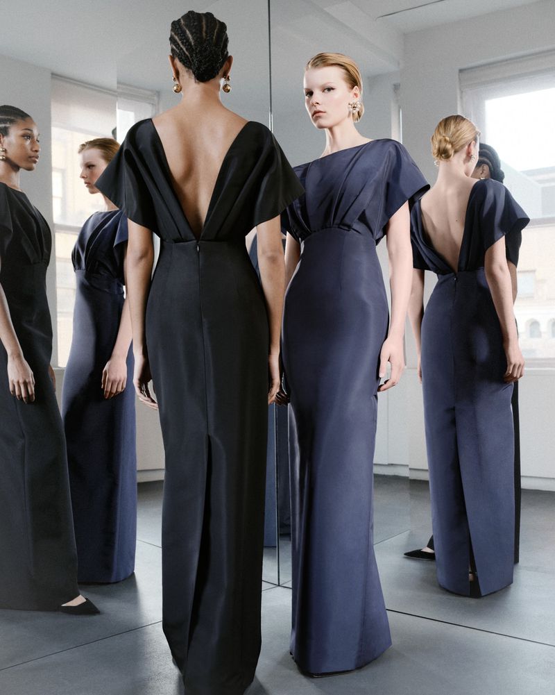 Five women in elegant, dark, formal gowns are seen in a brightly lit, neutral indoor setting with a mirror reflecting their images.