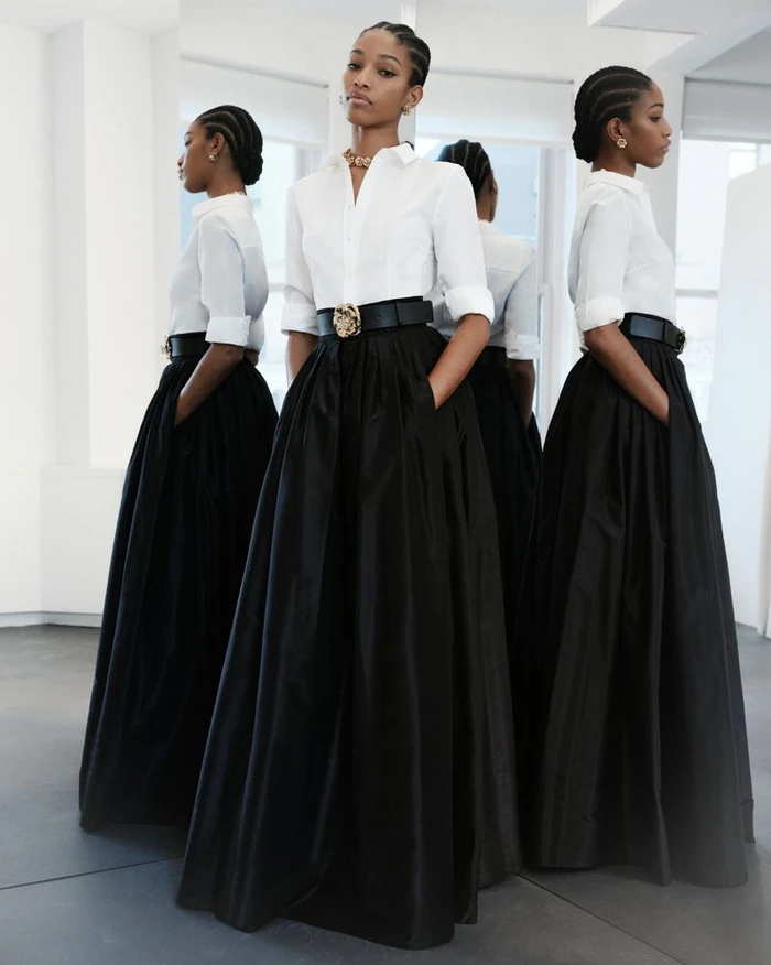 Model Naomi Chin Wing in a white button-down shirt and long black skirt standing before mirrors in a bright studio space