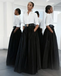 Model Naomi Chin Wing in a white button-down shirt and long black skirt standing before mirrors in a bright studio space