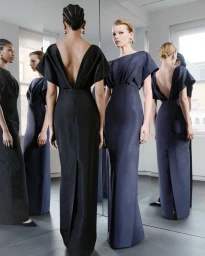 Two models in elegant black and navy floor-length gowns standing in front of mirrors in a sunlit studio space.