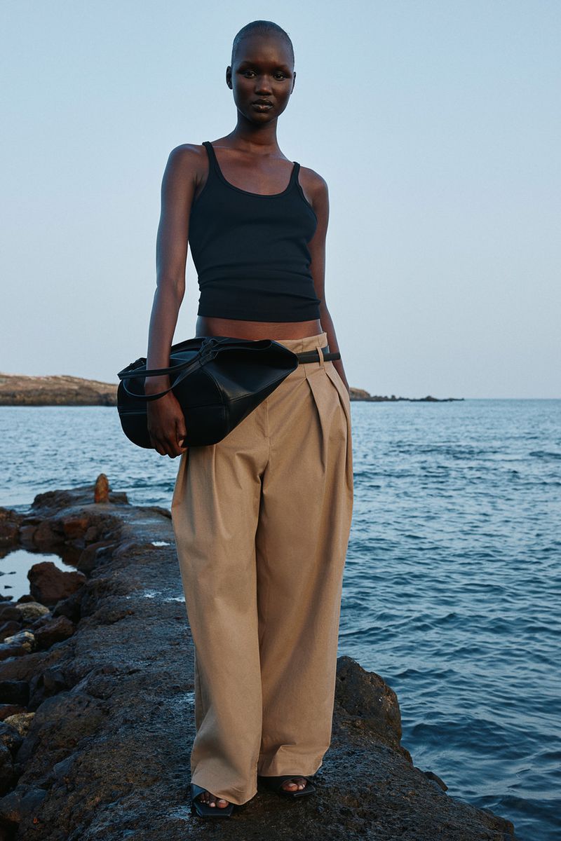 A solo woman stands on a stone pier by the sea, wearing a black tank top and beige trousers, accessorized with a dark bag and bracelet.