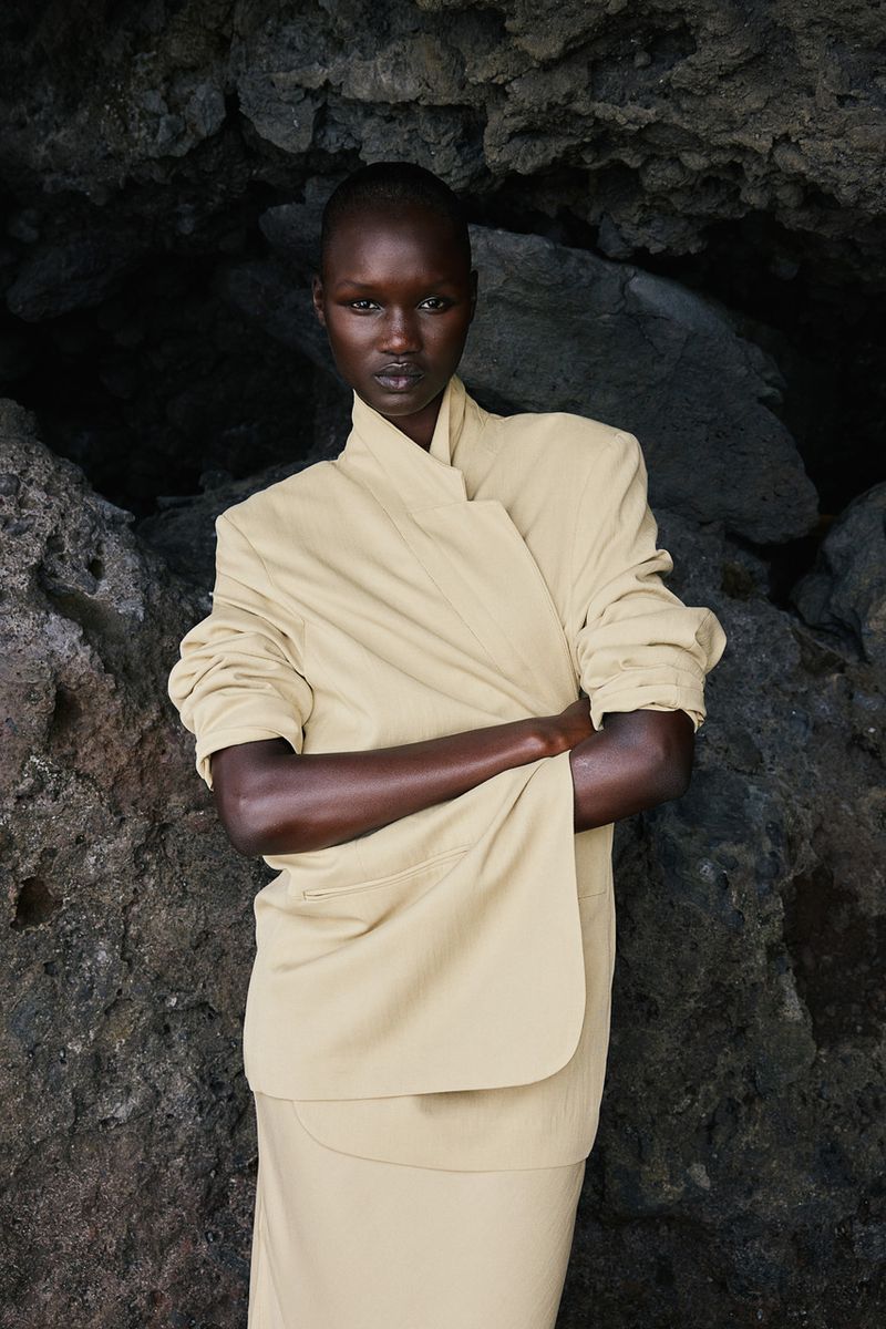 A striking portrait showcases a Black female model in a light beige blazer and skirt, posing with crossed arms against a dark stone background.