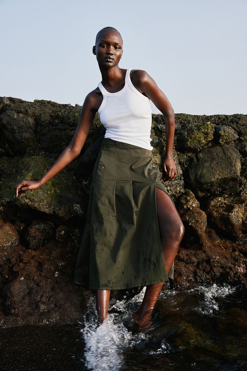 Fashion shot of an adult female wearing a tank top and skirt posing near water. The photo was taken outdoors with natural light.