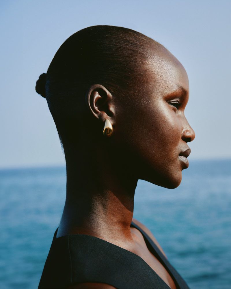 Close-up shot of a Black woman in profile wearing a black top and gold earrings against a blue sky and the sea, highlighting beauty and fashion.