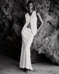 Black and white portrait of a female model standing against a textured rock formation wearing a buttoned vest and long skirt.
