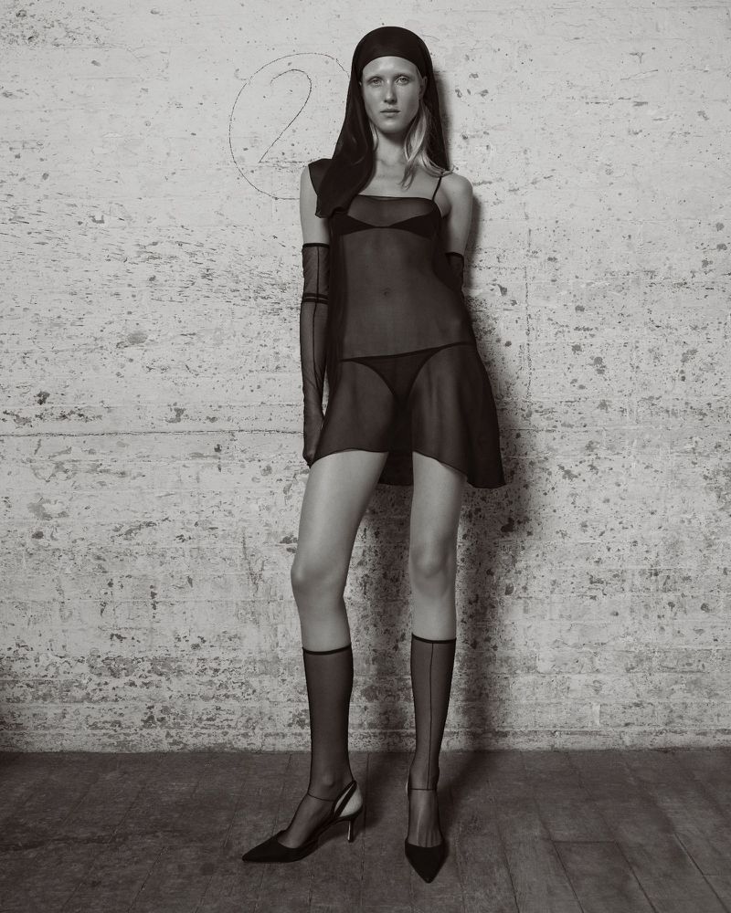 A slender woman poses in a sheer black dress, accessorized with opera gloves and knee-high socks against a textured gray wall backdrop.