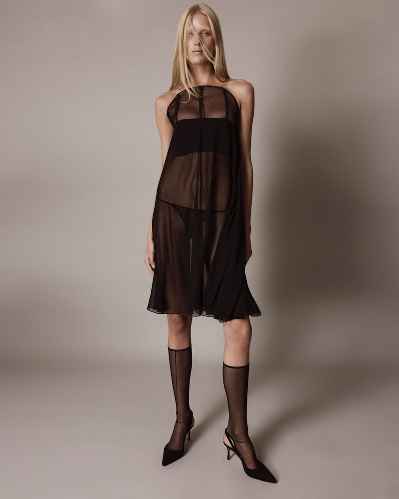 Elegant full-length shot of a blonde adult woman in a sheer black dress, stockings, and heels against a neutral grey studio backdrop.