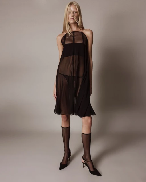 Elegant full-length shot of a blonde adult woman in a sheer black dress, stockings, and heels against a neutral grey studio backdrop.