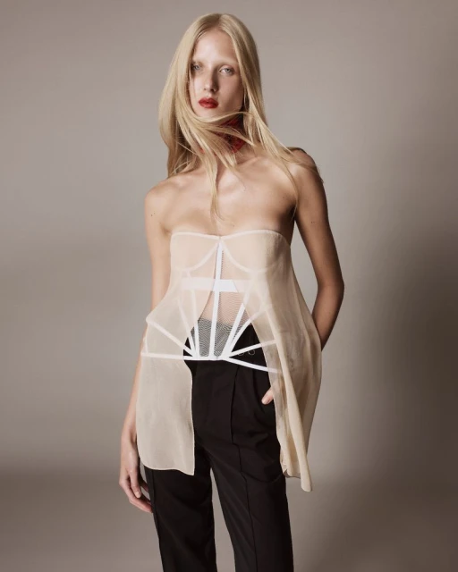 Fashion shot of a blonde woman wearing a sheer top and black pants, posing in a studio with a neutral background and red lipstick.