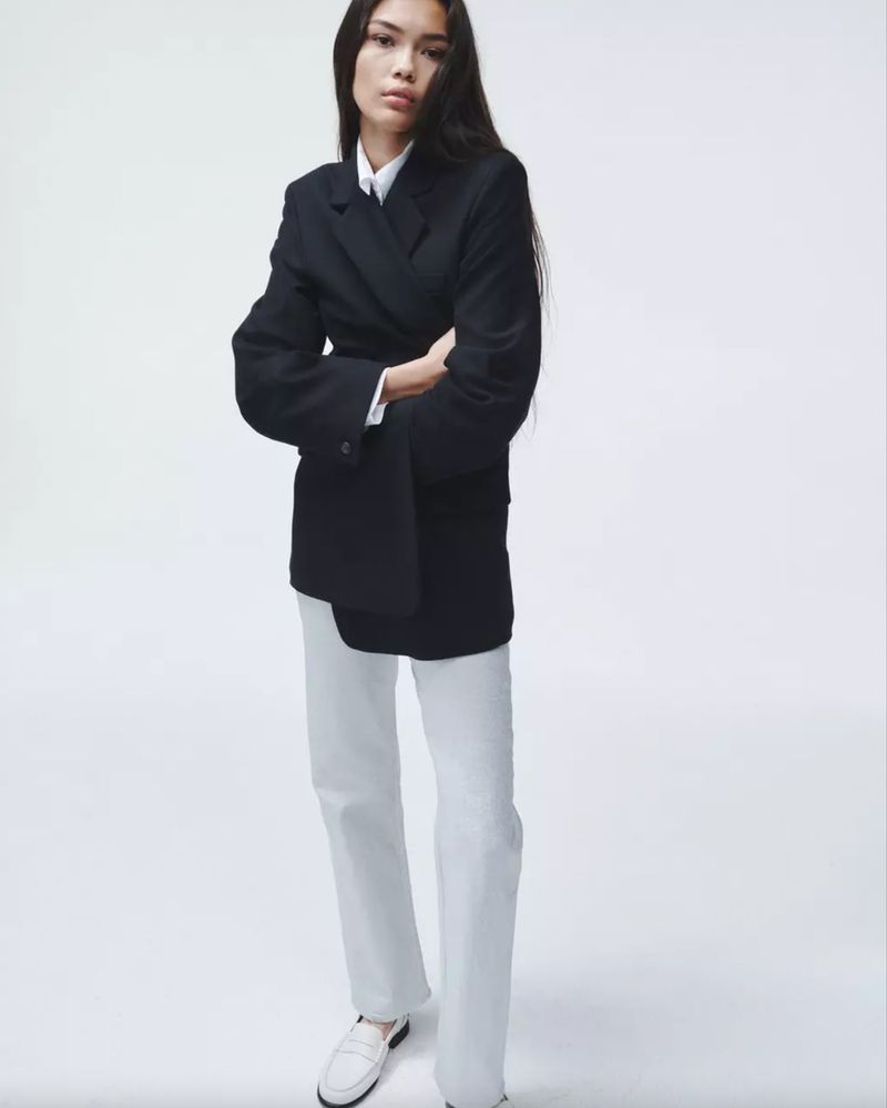 An adult female poses indoors wearing a double-breasted blazer, white top and pants, and loafers, set against a bright white backdrop.