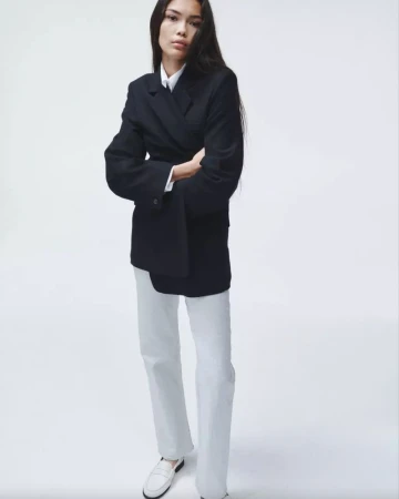 An adult female poses indoors wearing a double-breasted blazer, white top and pants, and loafers, set against a bright white backdrop.