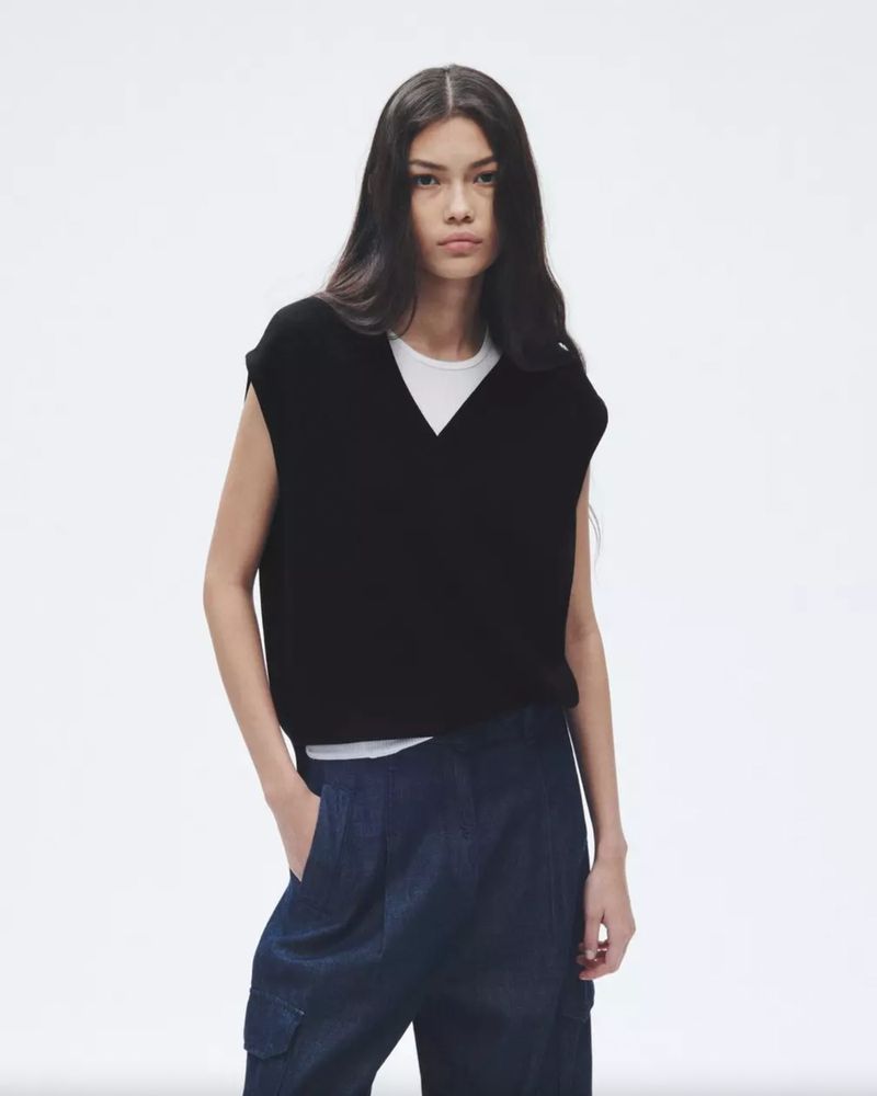 A woman with long, dark hair poses in a sleeveless black knit top and navy trousers against a stark white backdrop in a studio shot.