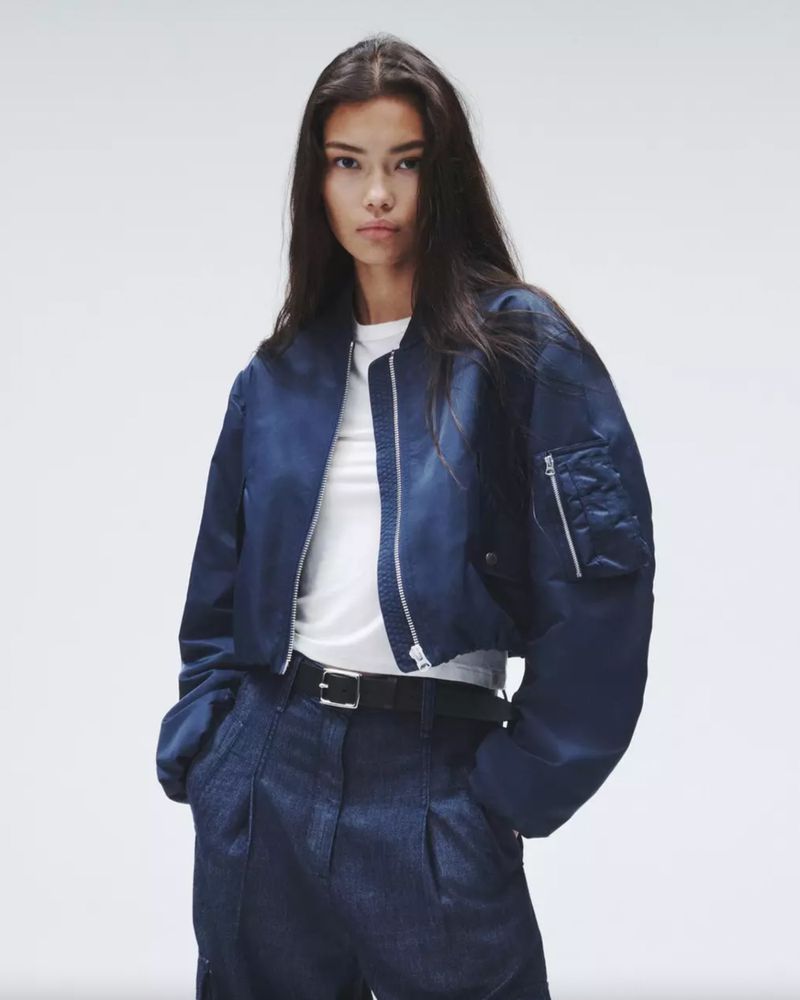 Young woman poses in a navy bomber jacket and matching trousers against a plain white backdrop in a studio fashion shot.