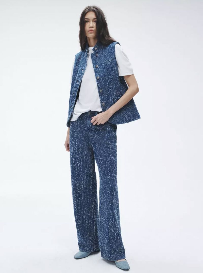 Full-shot of an adult woman in a white t-shirt, blue vest, and pants standing in a white studio setting. Fashion editorial photography.