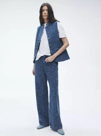 Full-shot of an adult woman in a white t-shirt, blue vest, and pants standing in a white studio setting. Fashion editorial photography.